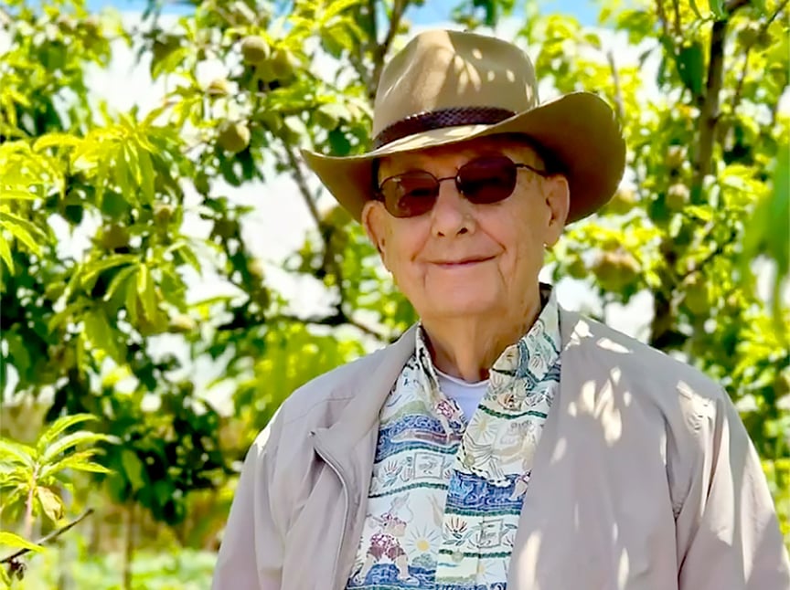 An elderly man wearing a tan hat, sunglasses, a patterned shirt, and a beige jacket stands smiling outdoors in front of green leafy trees on a sunny day.