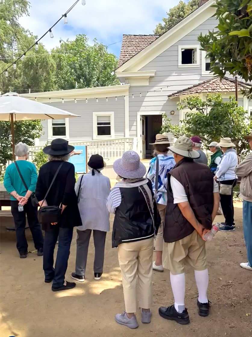 A group of people wearing hats and casual clothing stand outside a gray house with white trim, listening to a person speak. The scene is outdoors on a sunny day with trees and string lights visible.