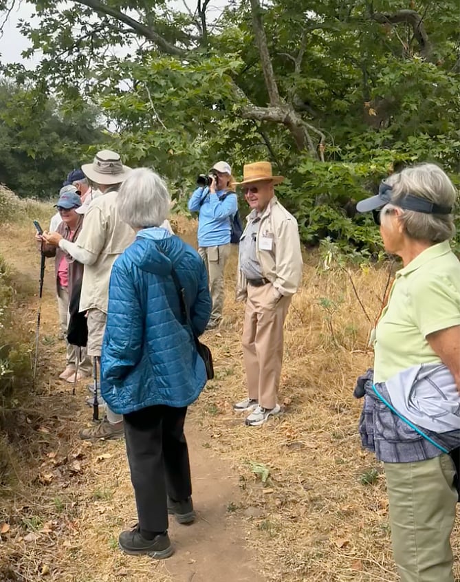 A group of people stand on a narrow dirt path in a grassy, wooded area. Some are wearing hats and jackets, and one person is taking photos with a camera. The group appears to be on a nature walk or hike.