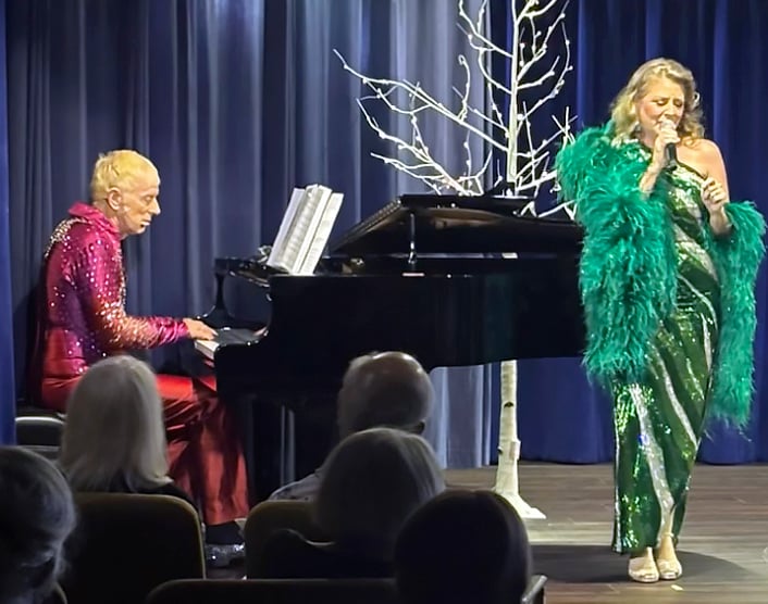 A man in a sparkly red outfit plays a grand piano while a woman in a green sequined dress and feathered shawl sings on stage; an audience watches in the foreground.