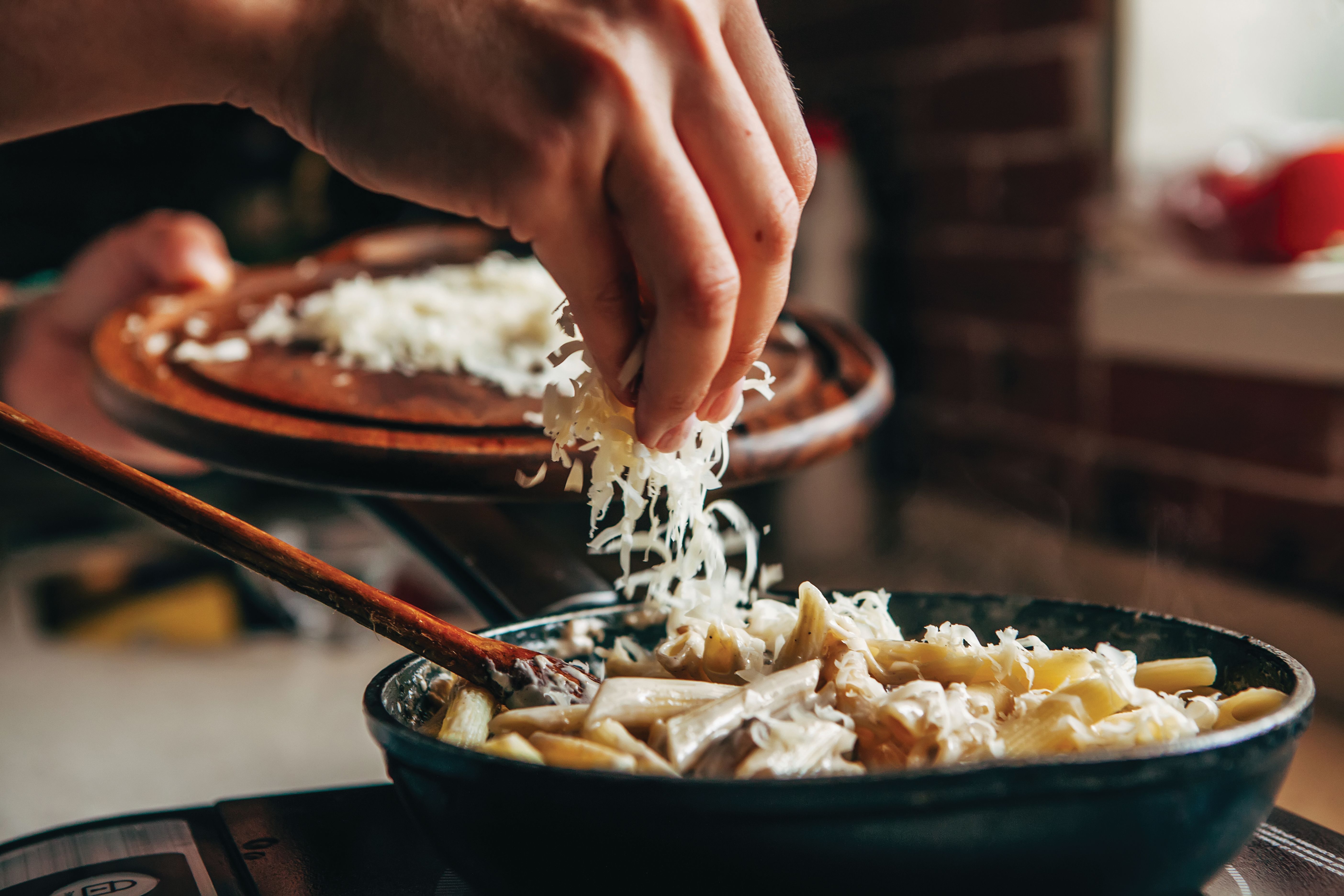 A close-up of a hand sprinkling grated cheese over a skillet filled with cooked penne pasta and sauce, with a wooden spoon resting in the pan.