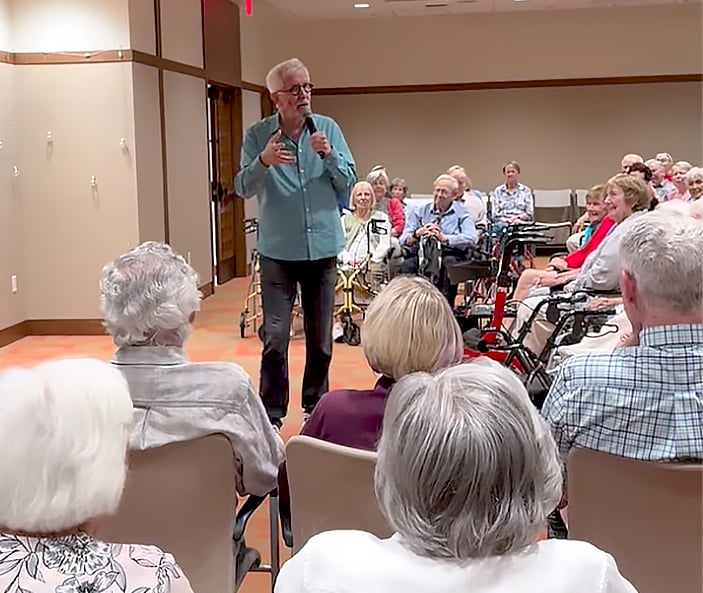 A man with gray hair and glasses speaks to a seated audience of elderly people in a community room. Several audience members use walkers, and all are attentively listening.