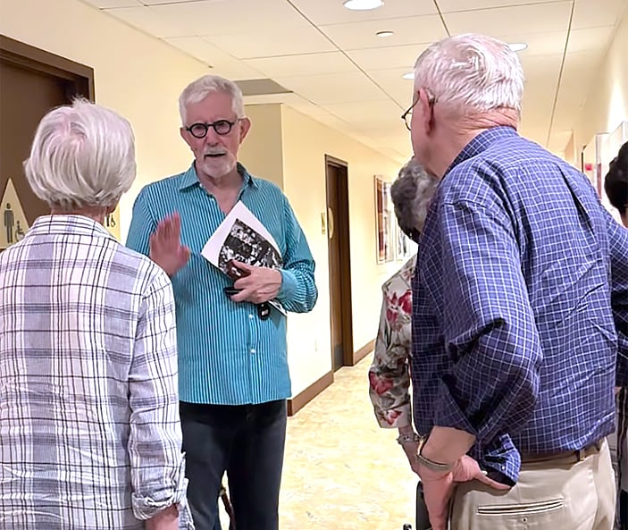 Four older adults stand and talk in a hallway. One man in a turquoise striped shirt holds a booklet and gestures while speaking to three others, two women and another man, all dressed in casual clothes.