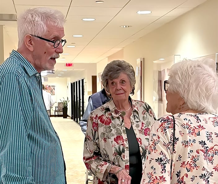 Three older adults, two women and one man, stand in a hallway having a conversation. All are wearing glasses and floral-patterned clothing. The hallway is brightly lit with beige walls and doors in the background.