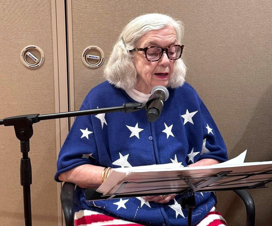 An older woman with white hair and glasses sits at a microphone, reading from papers. She is wearing a blue cardigan with white stars and a red-and-white striped skirt, resembling the American flag.