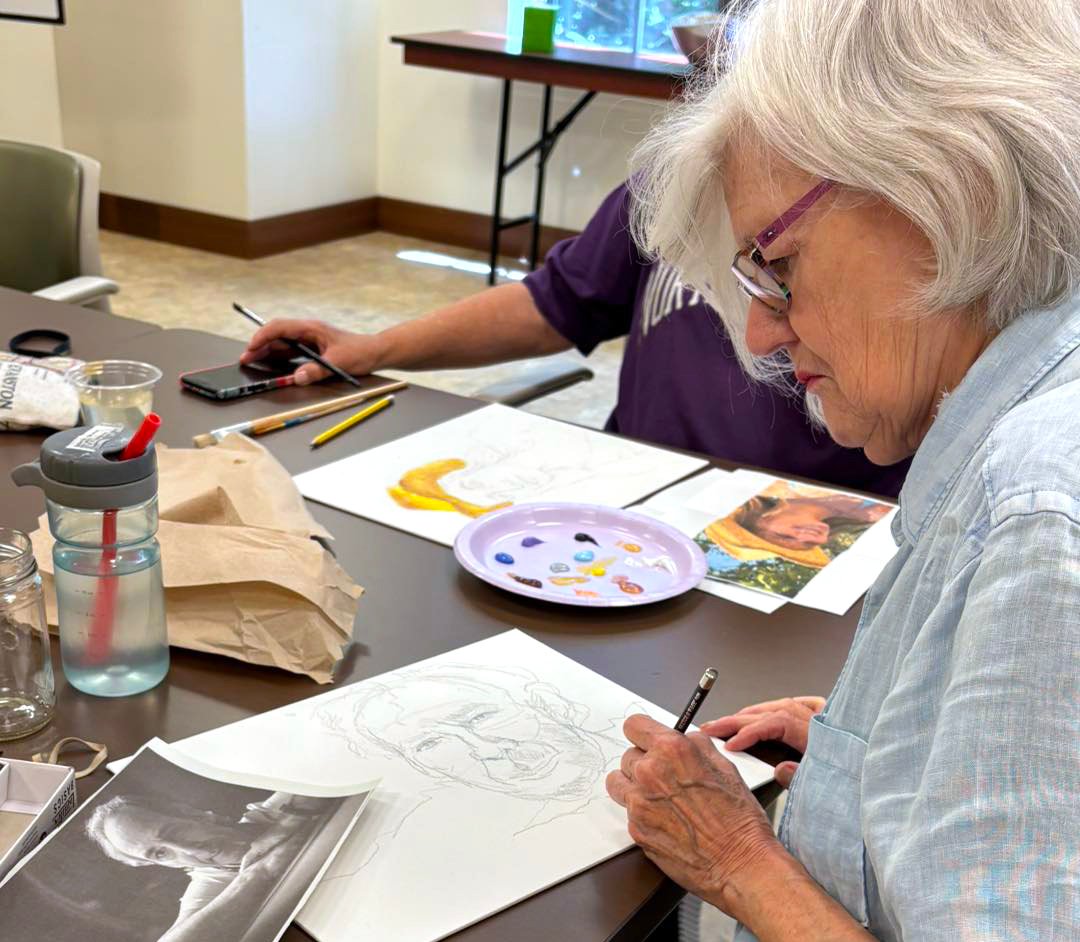 An elderly woman sketches a portrait with pencil at a table, referencing a printed photo. Art supplies, a palette, and sketches are scattered on the table. Another person’s hands are visible working nearby.
