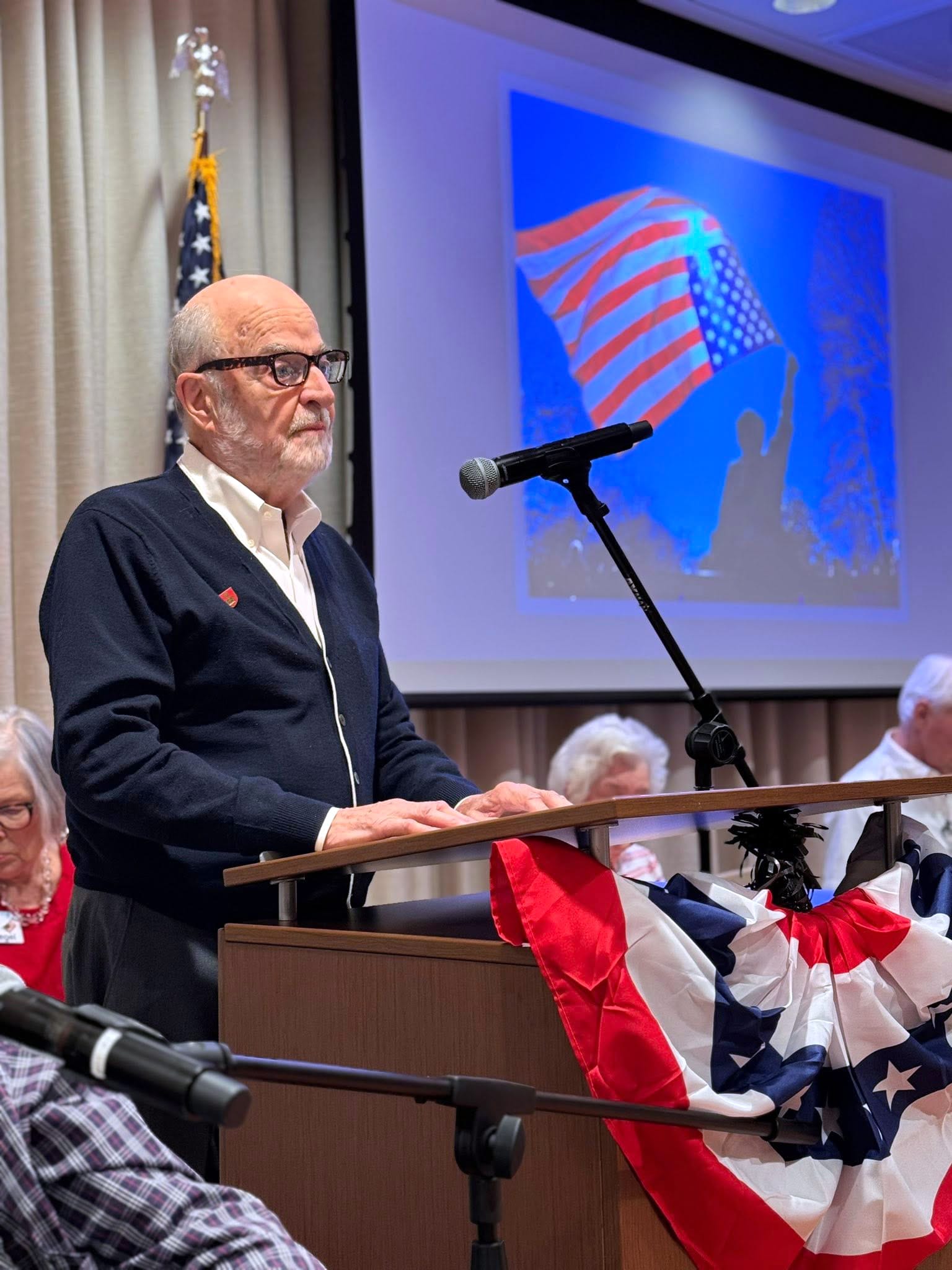 An older man stands at a podium decorated with patriotic red, white, and blue bunting, speaking into a microphone. Behind him is a screen showing an image of a waving American flag. Other people sit at a table nearby.