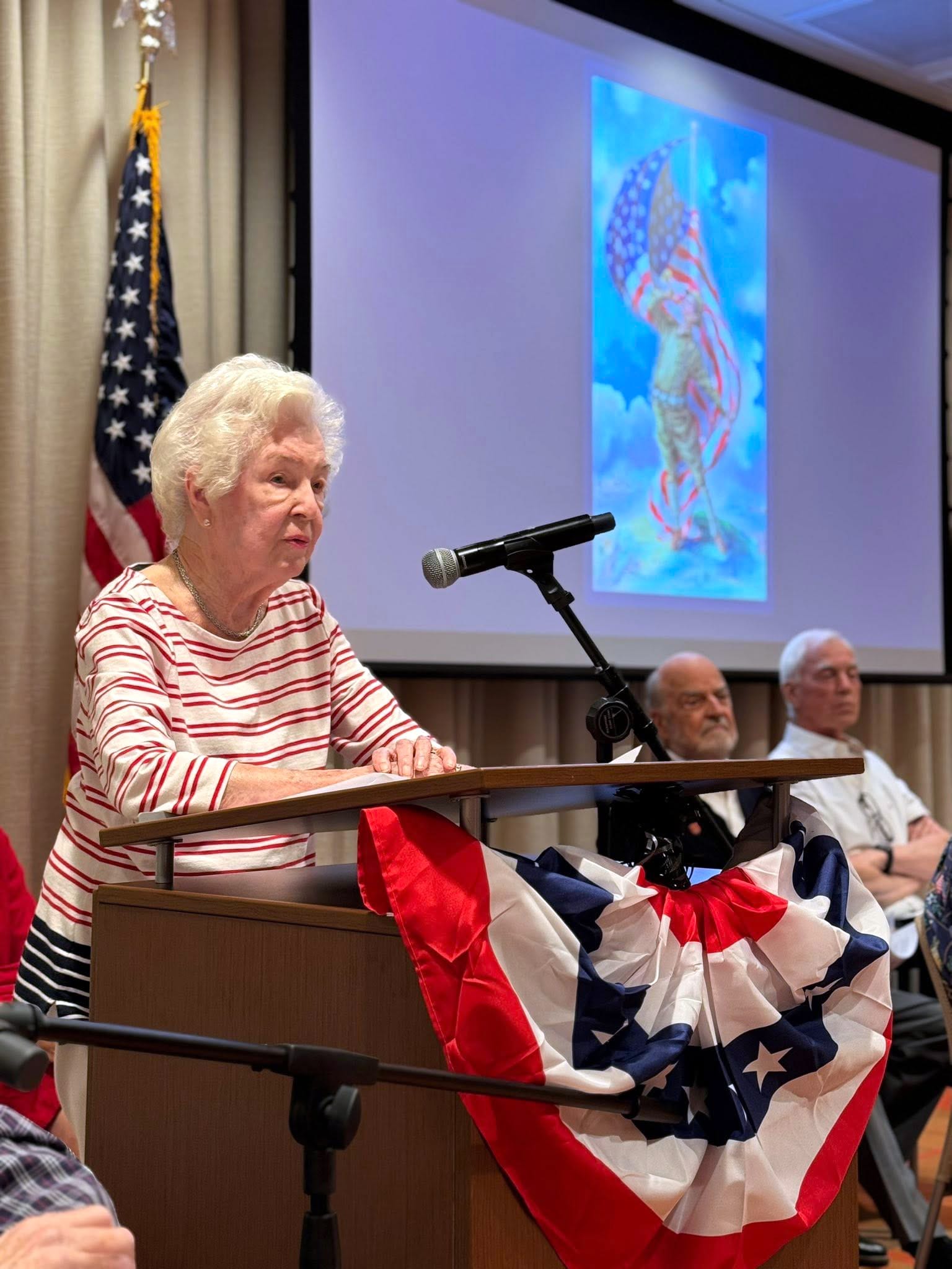 An elderly woman in a red and white striped shirt speaks at a podium decorated with a patriotic banner, with three older men seated behind her and an American flag and patriotic image displayed in the background.