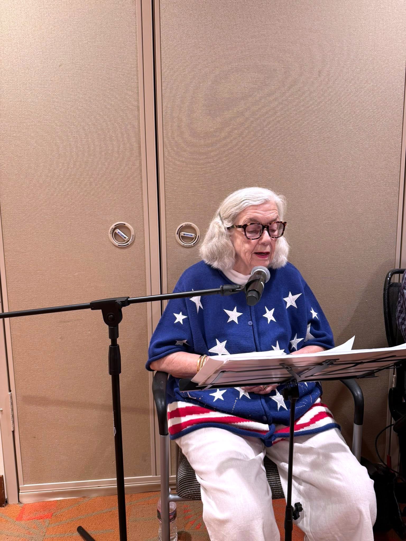 An elderly woman with white hair and glasses sits at a microphone, reading from papers. She wears a blue top with white stars and red stripes, resembling the American flag, and sits in front of beige folding doors.