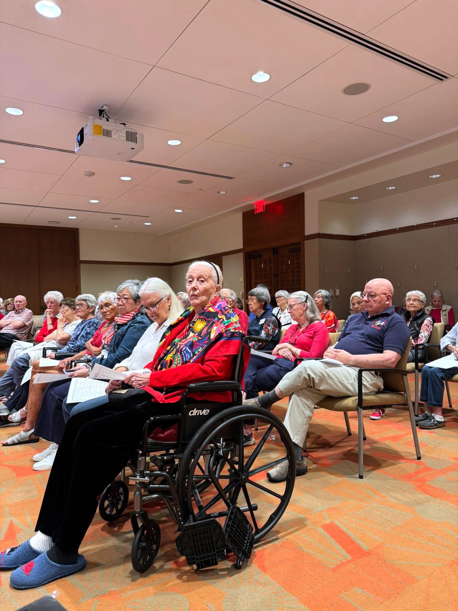 A group of elderly people sit attentively in a brightly lit room; a woman in a wheelchair with a colorful scarf sits at the front. The audience appears to be participating in a community or social event.