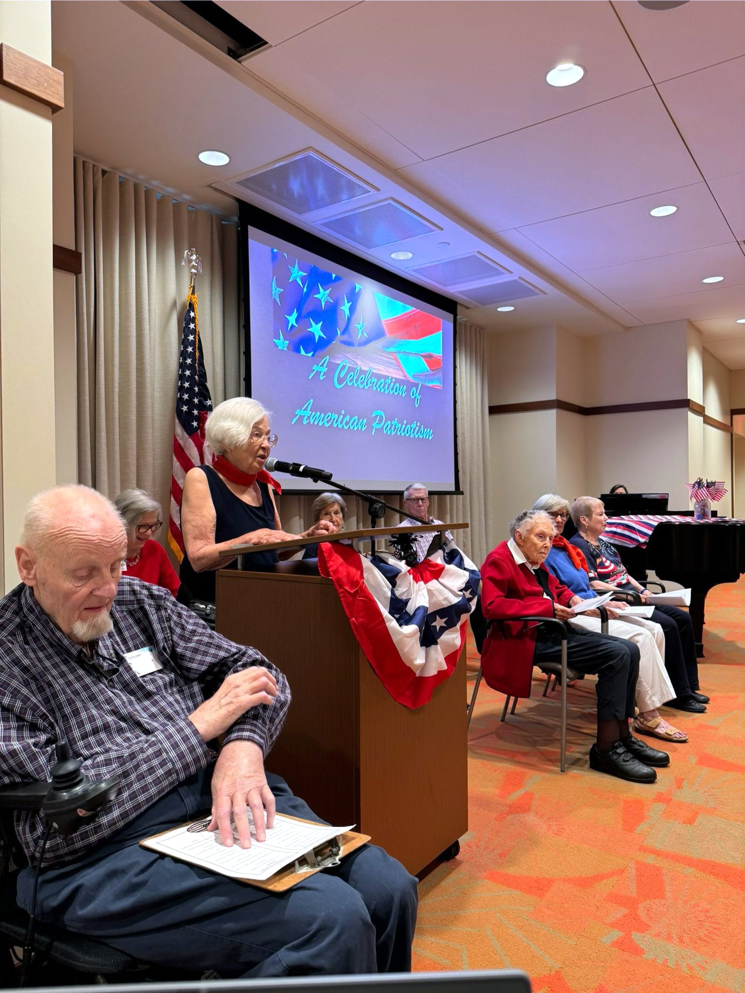 A woman speaks at a podium decorated with red, white, and blue bunting, while elderly people sit nearby. A screen behind displays A Celebration of American Patriotism with U.S. flag graphics.