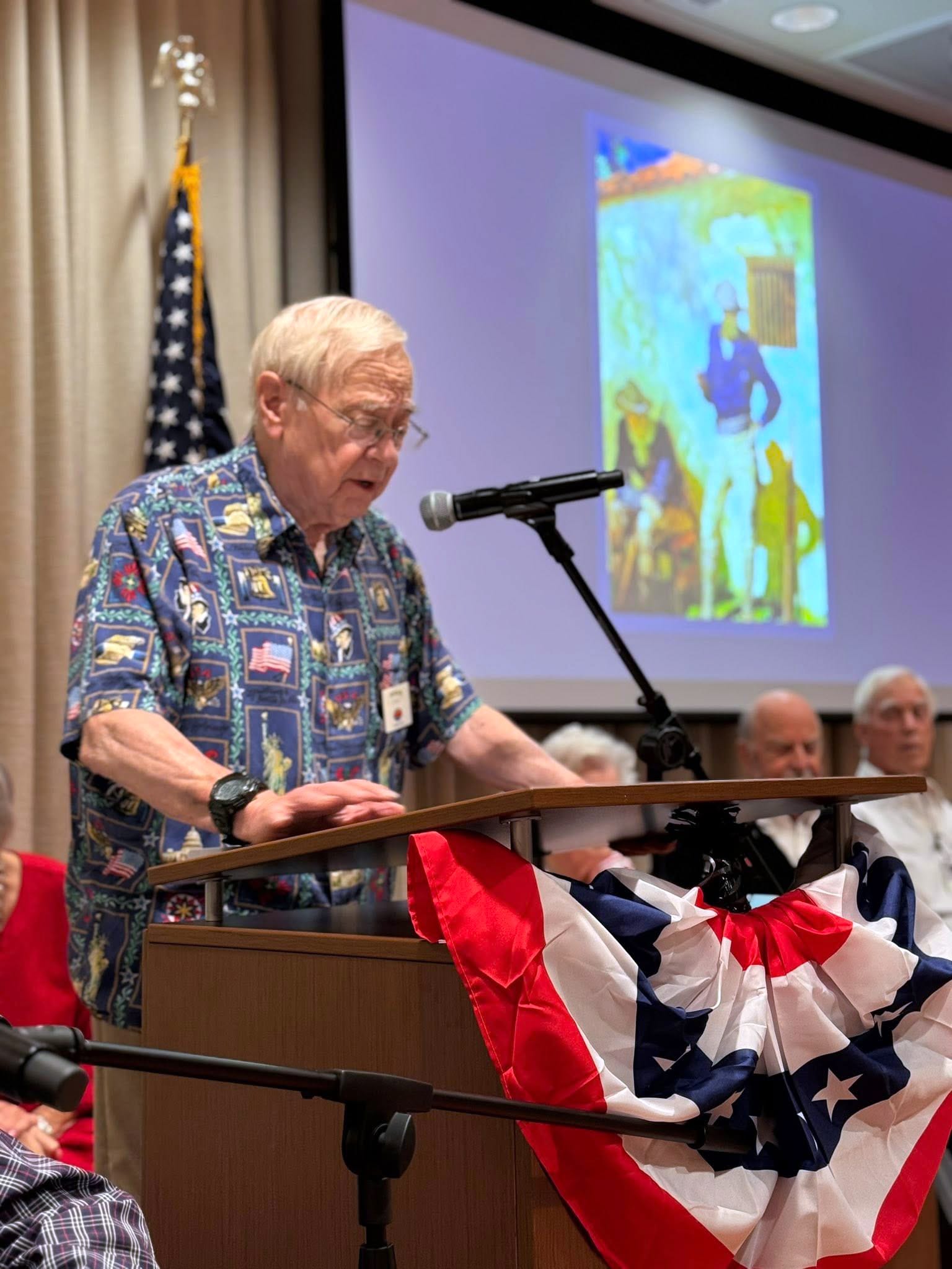 An elderly man in a patterned shirt speaks at a podium decorated with an American flag bunting, with an American flag and a projected image visible in the background. Several people are seated behind him.