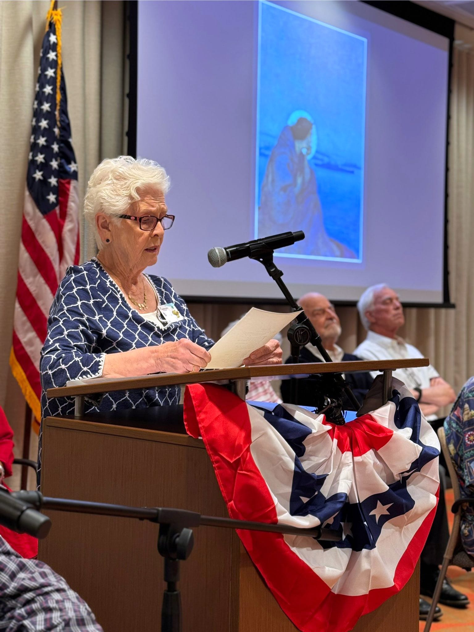 An elderly woman speaks at a podium decorated with patriotic bunting, with an American flag and three seated men in the background, and a blue-toned painting projected on a screen behind her.