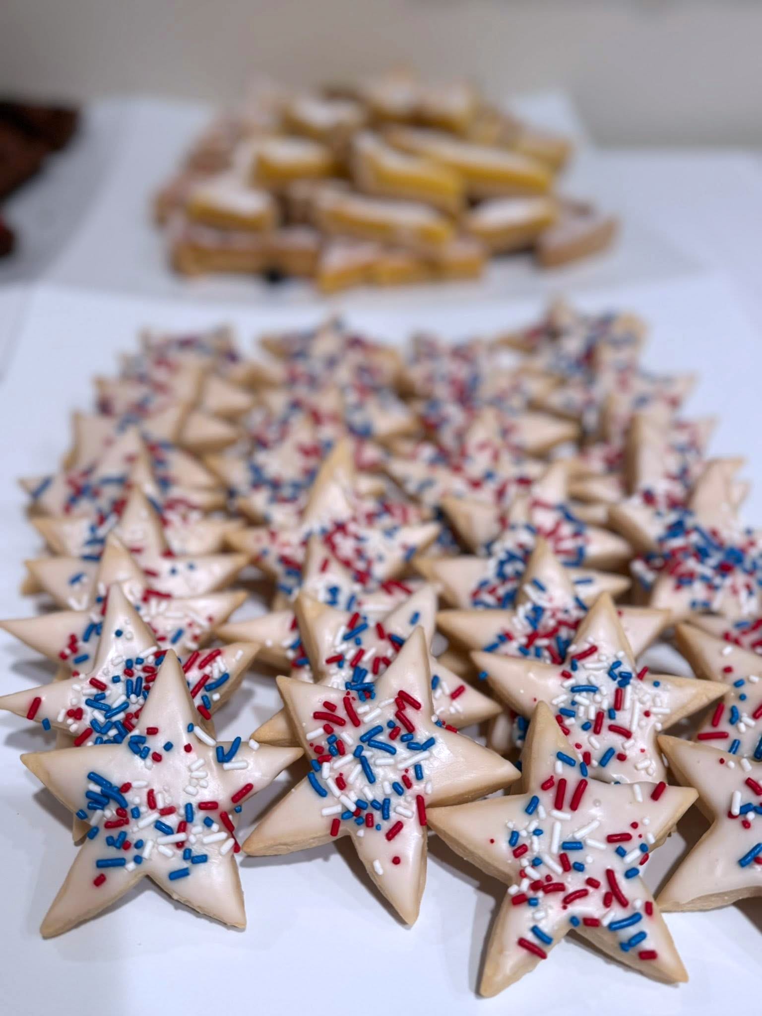 Star-shaped cookies with white icing and red, white, and blue sprinkles are arranged on a white surface. In the background, there are rectangular cookies with yellow icing stacked in a pile.