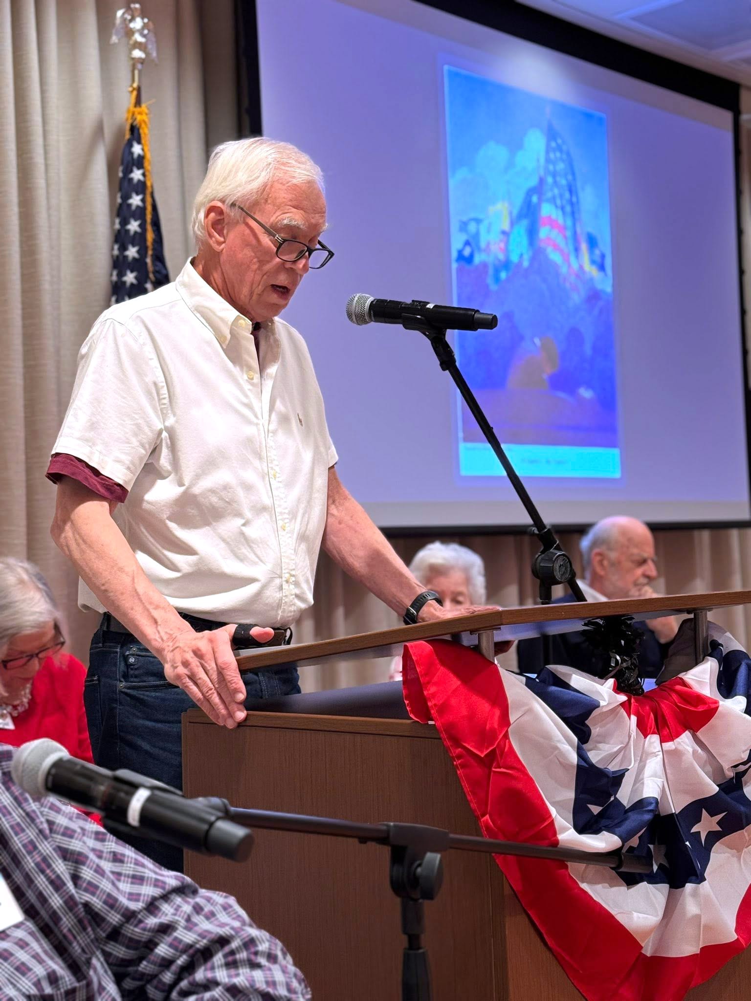An older man in a white shirt speaks at a podium decorated with a red, white, and blue banner. An American flag stands behind him, and several people are seated at a table. A projection screen displays an image in the background.