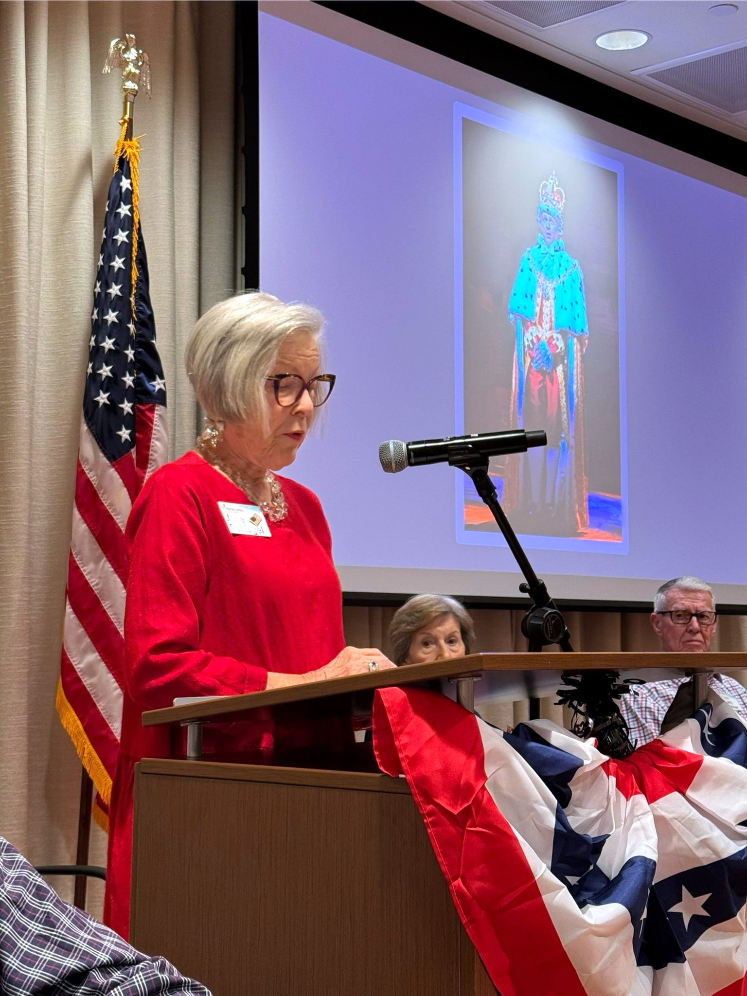 An older woman in a red dress speaks at a podium decorated with a patriotic bunting. An American flag stands behind her, and a large portrait of a crowned figure is projected on the screen. Several people sit nearby.