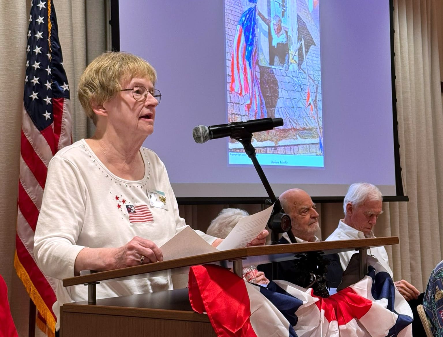 A woman speaking at a podium decorated with red, white, and blue bunting, with an American flag and elderly people seated behind her. A patriotic image is projected on the screen in the background.