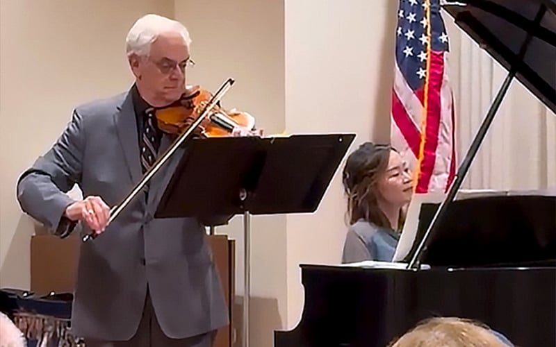 An older man in a suit plays the violin next to a woman playing a grand piano. An American flag is visible in the background inside a well-lit room.