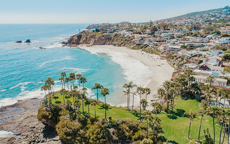 Aerial view of a coastal beach town with turquoise water, white sand, palm trees, and houses on hillsides overlooking the ocean under a clear blue sky.