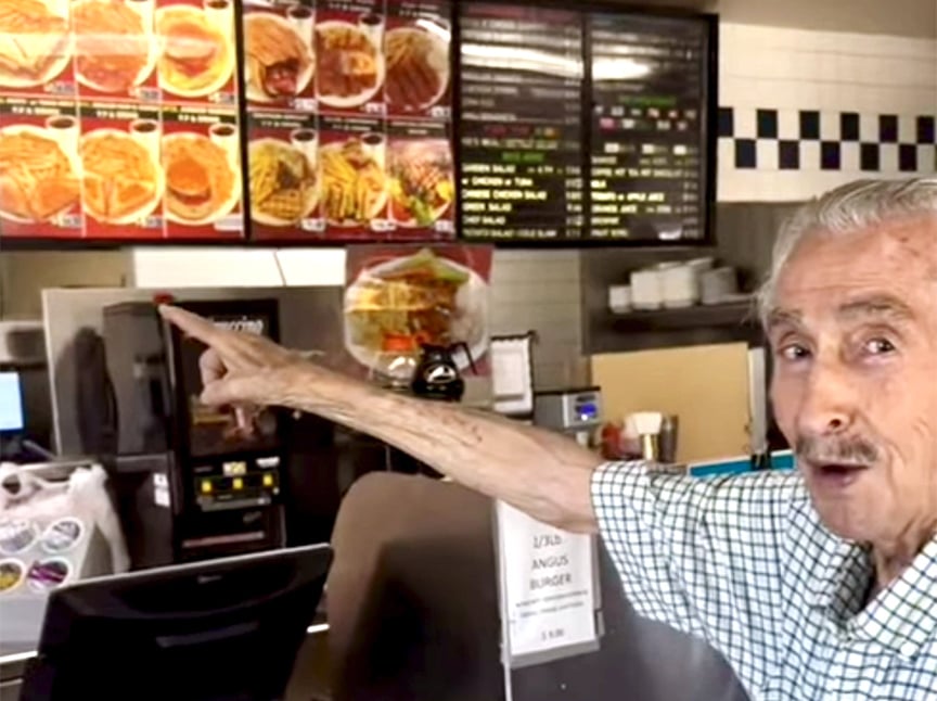 An elderly man in a checkered shirt points toward the menu behind the counter at a fast food restaurant, with burgers and fries pictured on the menu boards above.