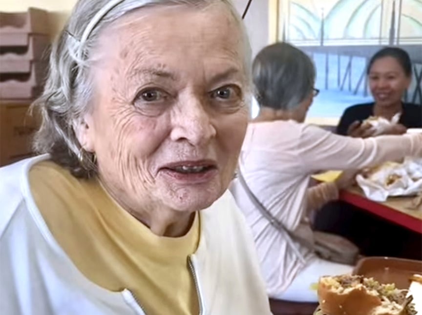 An older woman with gray hair and a yellow shirt smiles while holding a sandwich; two other women are seated at a table in the background, eating and chatting in a bright, casual setting.