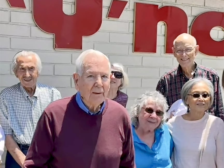 A group of six older adults, three men and three women, stand together and smile in front of a white brick wall with large red letters partially visible in the background.
