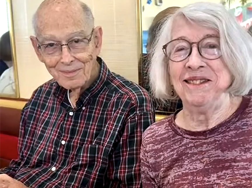 An older man in glasses and a plaid shirt sits next to an older woman with glasses and a patterned top. Both are smiling and sitting indoors, possibly at a restaurant.