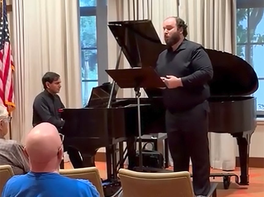 A man sings at a music stand beside a grand piano, while another man plays the piano. Several people sit and watch the performance in a room with tall windows and an American flag.