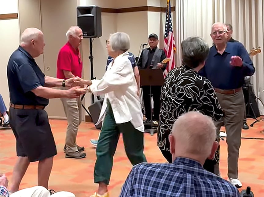 A group of older adults dance together in a brightly lit room while a small band performs in the background. The atmosphere is lively, with people smiling and enjoying the music. An American flag is visible by the stage.