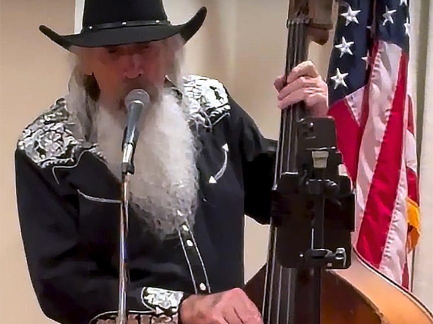 An older man with a long white beard, wearing a black cowboy hat and embroidered shirt, sings into a microphone while playing a double bass. An American flag is visible in the background.