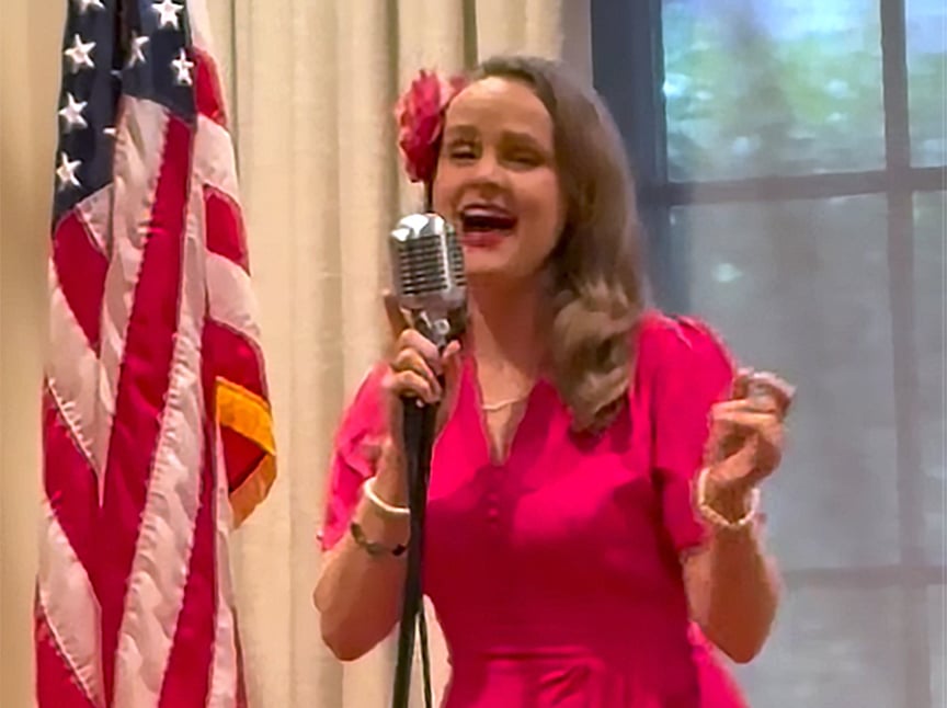 A woman in a bright pink dress sings into a vintage microphone next to a U.S. flag, with a flower in her hair and a large window in the background.