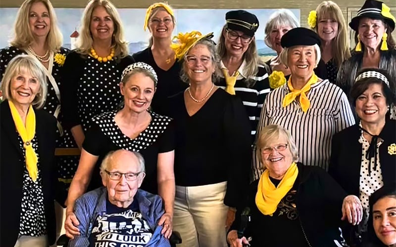 A group of smiling women and one elderly man pose together indoors. Many are wearing black and white outfits with yellow accents such as scarves and flowers. The setting looks festive and cheerful.