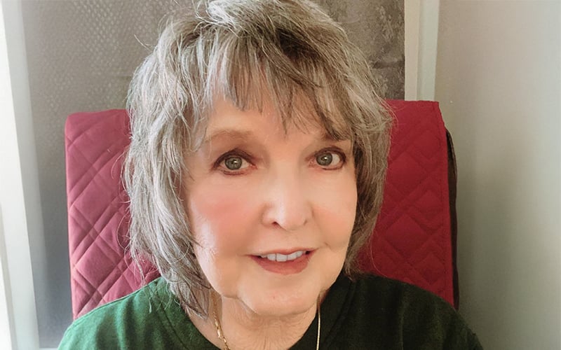 A smiling older woman with short, gray hair sits indoors in front of a red, quilted background, wearing a green top.