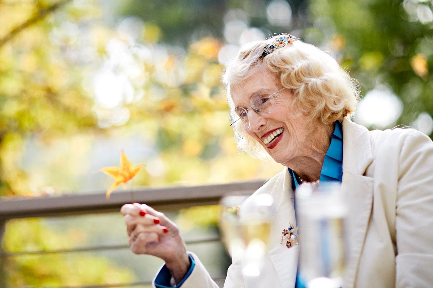 An elderly woman with blond hair and glasses smiles while holding a yellow autumn leaf. She is wearing a cream jacket and blue shirt, sitting outdoors with blurred greenery and drinks in the foreground.
