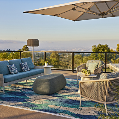 Outdoor patio with a blue sofa, cushioned chair, modern coffee tables, and a large umbrella on a patterned rug. Trees, greenery, and a distant view under a clear sky are in the background.