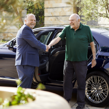Two older men smile and shake hands beside a black car; one wears a suit, the other a green polo shirt and dark pants. The car door is open and trees are visible in the background.