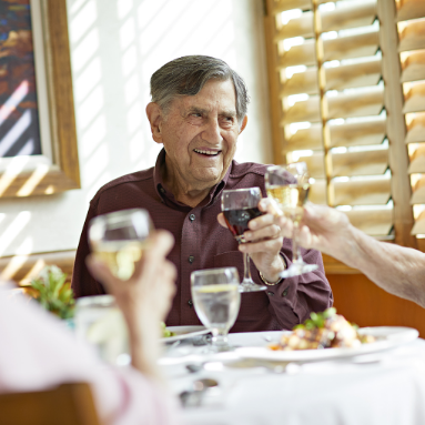 An elderly man smiles while raising a glass of red wine in a toast with friends at a sunlit restaurant table set with water, wine, and plates of food.