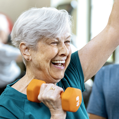 Smiling elderly woman with short gray hair lifts an orange 5 lb dumbbell while exercising indoors, wearing a teal shirt.