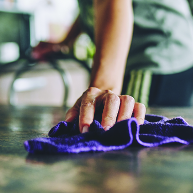 A person’s hand cleaning a surface with a purple cloth, while holding onto a chair in the background.