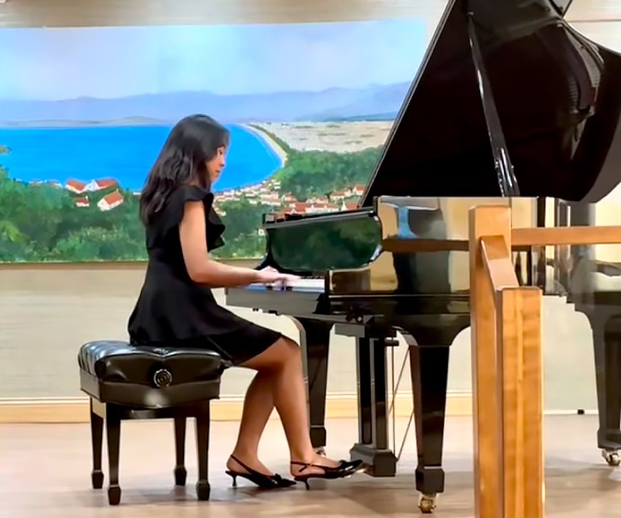 A young woman in a black dress plays a grand piano on stage. Behind her, there is a colorful painting of a coastal landscape with blue water, green hills, and buildings.