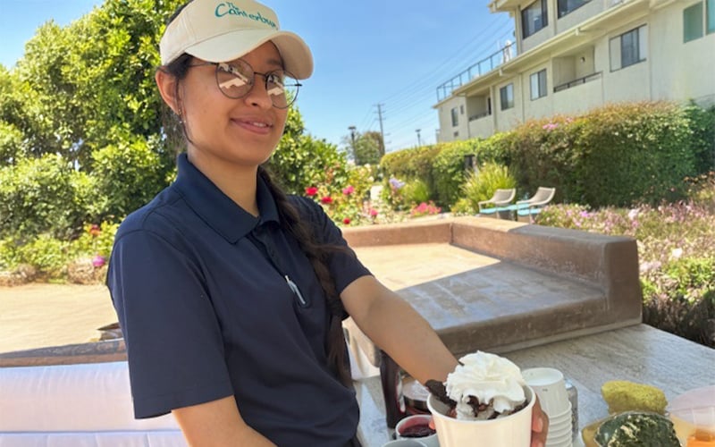 A woman wearing a beige hat and navy shirt smiles while serving a dessert with whipped cream outdoors, with plants, flowers, and buildings in the background.