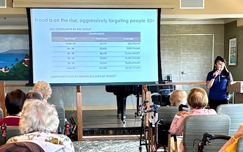 A woman gives a presentation to a group of older adults. A slide on the screen reads Fraud is on the rise, aggressively targeting people 50+, with a table showing fraud complaints by age group.
