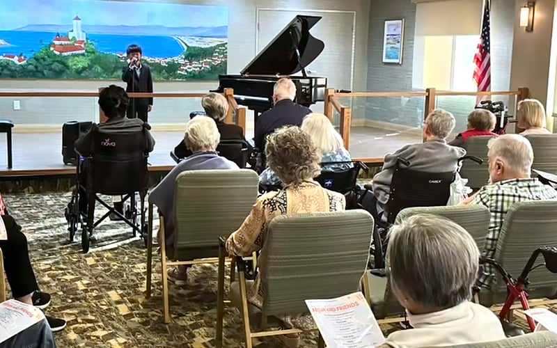A group of elderly people seated in a community room watch a performer singing at a microphone beside a grand piano, with an ocean landscape mural and an American flag in the background.