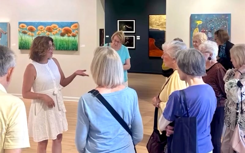 A woman in a white dress speaks to a group of older adults in an art gallery. Colorful paintings hang on the walls behind them. The group listens attentively as she gestures with her hand.