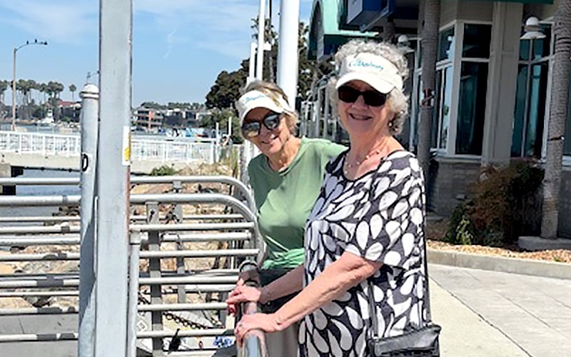 Two older women wearing sunglasses and visors stand smiling by a metal railing outdoors near a waterside walkway, with buildings and trees in the background on a sunny day.