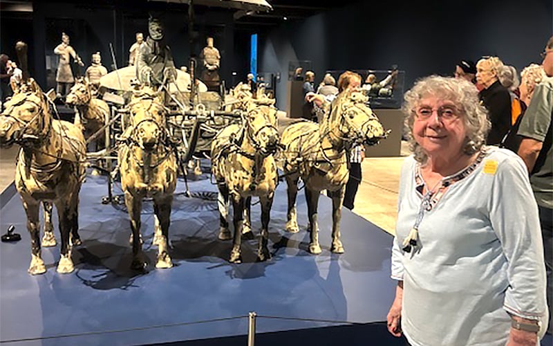 An older woman smiles in front of a display featuring life-sized terracotta horses pulling a chariot, with terracotta warrior statues visible in the background at a museum exhibit.