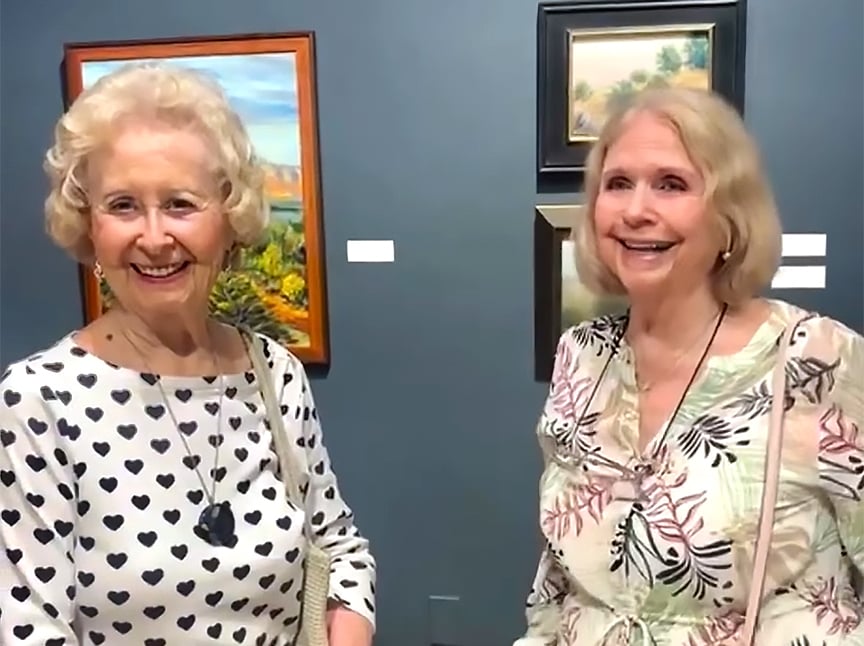 Two older women smiling and standing together in an art gallery, with colorful paintings hanging on the blue wall behind them. One woman wears a white top with black hearts; the other wears a floral blouse.