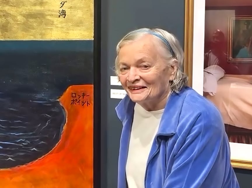 An elderly woman with gray hair, wearing a blue jacket and white shirt, stands smiling next to a colorful painting featuring a dark sea and orange shore. Framed artwork is visible in the background.