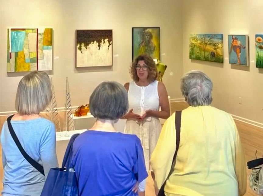 A woman in a white dress speaks to three women in a gallery, surrounded by colorful paintings on cream-colored walls. The listeners have gray hair and wear blue and yellow tops.