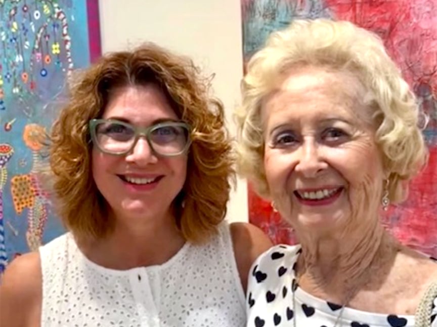 Two smiling women stand close together in front of colorful abstract art. One has curly light brown hair and glasses, and the other has short, curly blonde hair and is wearing a white top with black spots.